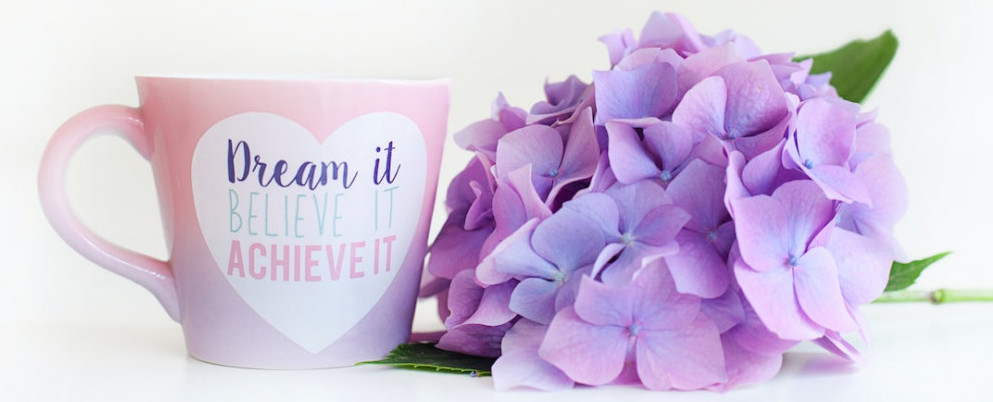 A pink coffee cup next to lavender flowers.  Coffee cup displays words dream it, believe it, achieve it. 